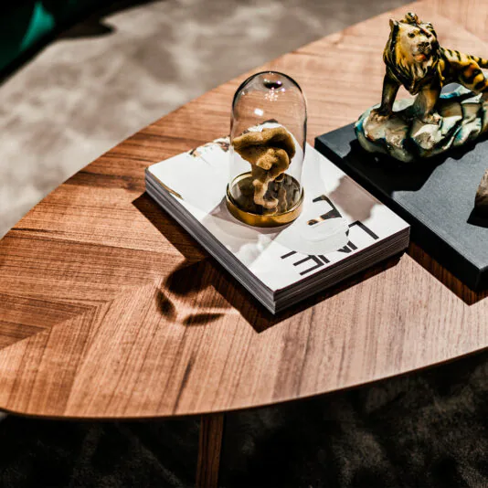 Large Designer Walnut Oval Coffee Table A Large Designer Walnut Oval Coffee Table holds a stack of books, with one topped by a mushroom encased in glass and adorned with a ceramic lion sculpture. The table is placed on a dark carpeted floor, and part of a green sofa is visible in the background.