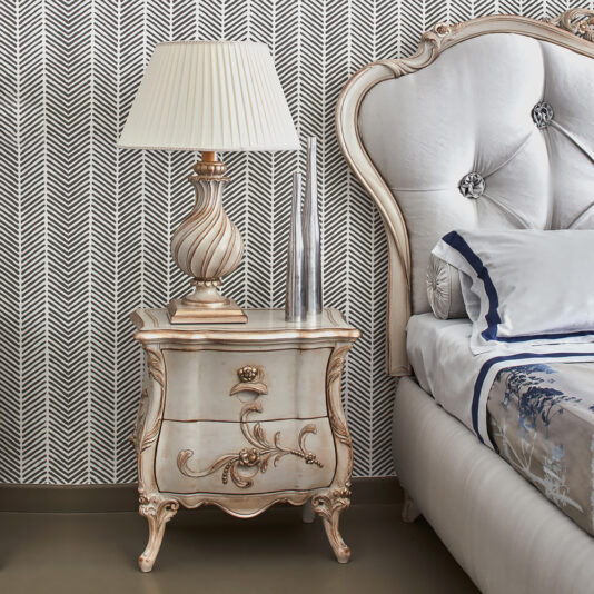 Elegant bedroom with a bed featuring a tufted headboard and plush bedding. A decorative lamp and metallic vase sit on classic Italian designer bombe bedside drawers. The wall showcases a black and white herringbone pattern.