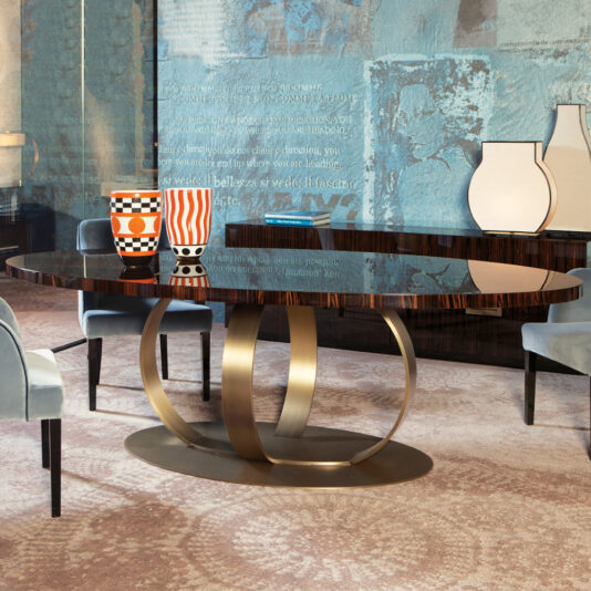 A modern dining room features a sleek metallic base Contemporary Italian bronzed ring oval dining table adorned with two geometric-patterned orange and black vases, surrounded by light blue upholstered chairs and accented with abstract wall art.