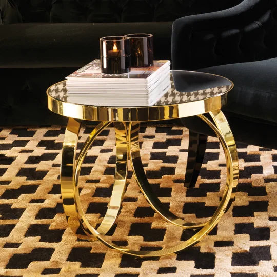 The High End Italian Brass Ring Side Table, featuring a glossy black top and a looped base design, holds two black candles and books. It stands elegantly on a patterned brown and black carpet beside a sleek black upholstered chair.
