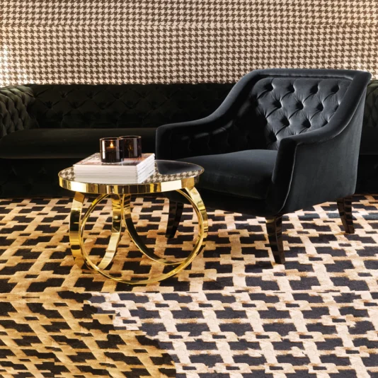 High End Italian Brass Ring Side Table A stylish room showcases a black tufted armchair and sofa against a houndstooth-pattern wall. A modern round gold table with mirrored top holds books and candles, complemented by the High End Italian Brass Ring Side Table. The floor features a matching houndstooth-pattern rug.