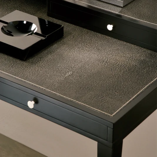 The Modern Italian Leather Writing Desk features a black textured surface with white knobs on its drawers. Atop the desk, a glossy black ashtray and matching square object reflect light over its subtly speckled pattern.