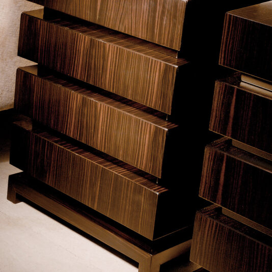 Close-up of two Luxury Ebony Four Drawer Chests, featuring a sleek, angular design with a dark, glossy finish. The slightly open drawers reveal rich wood grain patterns. These elegant pieces stand on hardwood flooring against a textured wall.