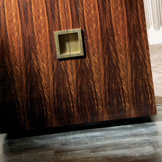 A close-up of the Italian Designer High End Ebony Veneer Cabinet features a square metallic handle on a polished door. The rich, dark brown finish exudes luxury, complemented by a light gray floor that highlights the exquisite craftsmanship.
