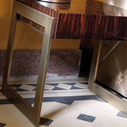 High End Contemporary Macassar Bedside Table A modern desk with a glossy wooden surface and bronze legs stands on a patterned tile floor. A tufted chair is partially visible beside the desk. The wall behind features an earthy, textured finish reminiscent of the High End Contemporary Macassar Bedside Table.