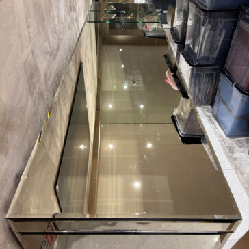 Luxury High End Wall Mounted TV and Media Unit Set A mirrored console table reflects ceiling lights and the room, complementing the Luxury High-End Wall-Mounted TV and Media Unit Set. Black storage bins are stacked to the right, against a beige wall and tiled floor.
