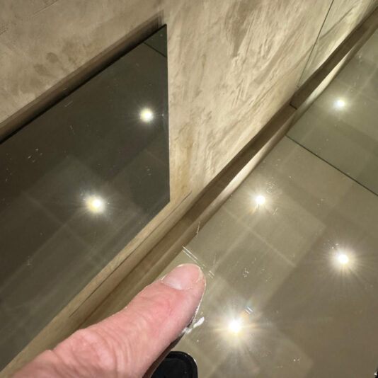 A finger points at a scratch on reflective glass, with bright ceiling lights and a textured beige wall behind, near the Luxury High-End Wall-Mounted TV and Media Unit Set.