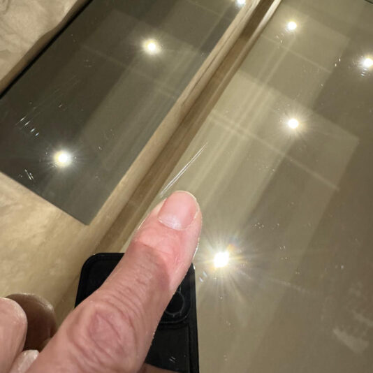 A hand points to a visible scratch on the reflective glass of the Luxury High-End Wall-Mounted TV and Media Unit Set, with bright ceiling lights reflected on the surface.