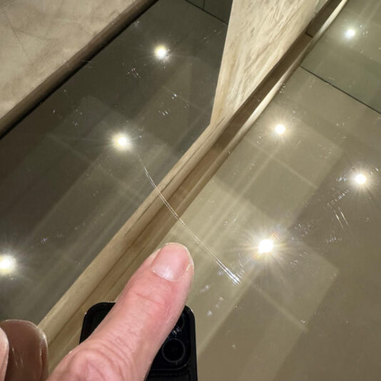 A finger points at a thin scratch on the reflective glass of the Luxury High-End Wall-Mounted TV and Media Unit Set, with bright ceiling lights mirrored in the background.