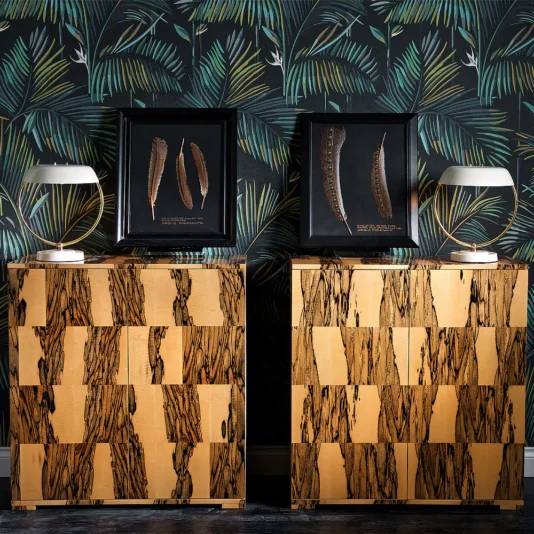 Two Luxury Italian Albin wood veneered cabinets with a checkered pattern are set against tropical leaf-patterned wallpaper, each topped with a modern white lamp and a framed decorative feather display.