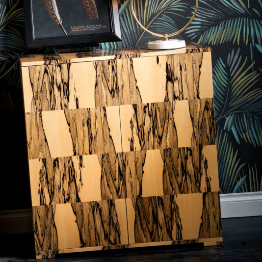 A Luxury Italian Albin Wood Veneered Cabinet with a checkered light and dark wood pattern stands against a leafy wall. On top, it features a circular gold-toned lamp and a picture frame displaying a feather design.