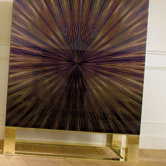 Modern High End Italian Ebony Veneered Cabinet The Modern High End Italian Ebony Veneered Cabinet features a starburst front pattern and luxe wood grain in brown, purple, and ebony. It's elegantly perched on a gold metallic base against a white wall and wooden floor.