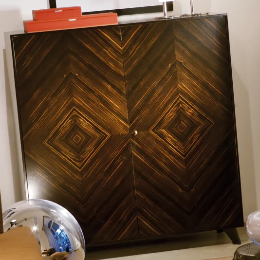 Luxury Italian Patterned Ebony Veneered Cabinet The Luxury Italian Patterned Ebony Veneered Cabinet features intricate diamond patterns in rich ebony wood, creating a symmetrical grain design. On top are a small decoration and red box, while a reflective metallic object is partially visible in the foreground.