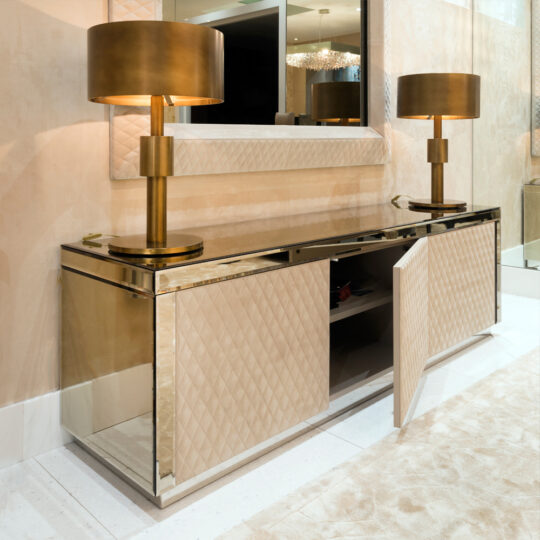 Luxurious interior showcasing a mirrored sideboard with quilted beige panels and an open door, flanked by two metallic gold lamps with drum shades. A large textured-frame mirror hangs above, complemented by a plush beige carpet and the Luxury High-End Wall Mounted TV and Media Unit Set.
