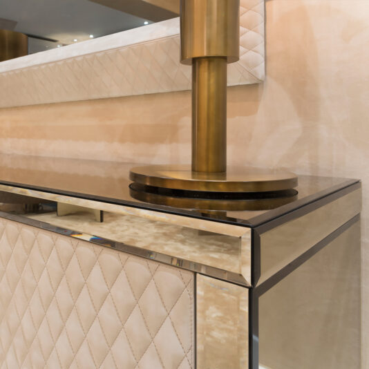 The modern interior design showcases a quilted beige wall panel, glass-topped console, and brass lamp with a cylindrical base. These elegant textures and metallic finishes pair beautifully with the Luxury High End Wall Mounted TV and Media Unit Set for a luxurious contemporary look.