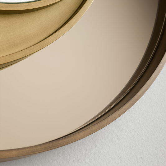 Close-up of the Modern Italian Designer Offset Circles Mirror, which showcases an offset circular design with a gold-toned frame at the top edge and a muted gray frame towards the bottom. This designer mirror reflects a minimalistic interior and is mounted on a white-textured wall, exemplifying sleek contemporary elegance.