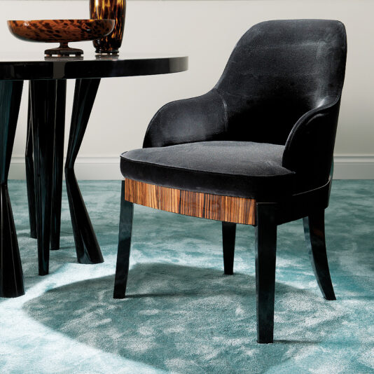 The Designer Italian Macassar Velvet Dining Chair, with its elegant curved armrests and cushioned back, sits on a light blue carpet beside a round black table, adorned with wooden accents. The table holds a decorative bowl.