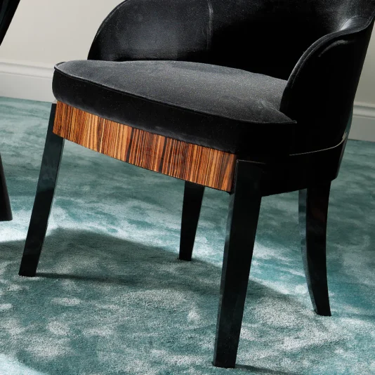 The Designer Italian Macassar Velvet Dining Chair showcases curved armrests, four angled legs, and a wooden Macassar accent on the front edge in sleek black, all elegantly positioned on a soft teal carpet.