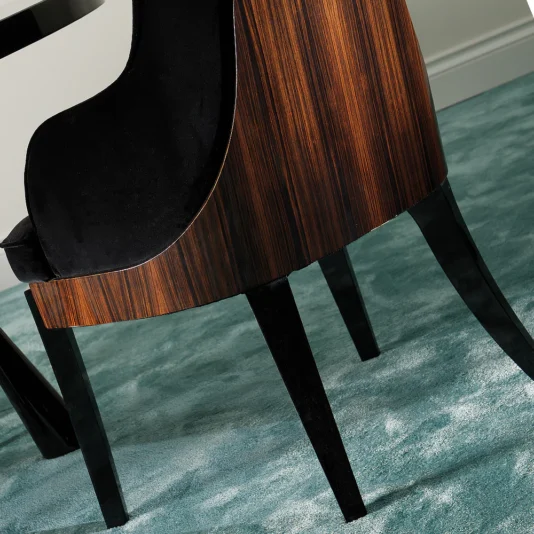 The Designer Italian Macassar Velvet Dining Chair, featuring a curved wooden backrest and elegant black upholstery, rests on a light teal carpet. Its black legs complement the luxurious design, while a corner of a refined black table is visible beside it.