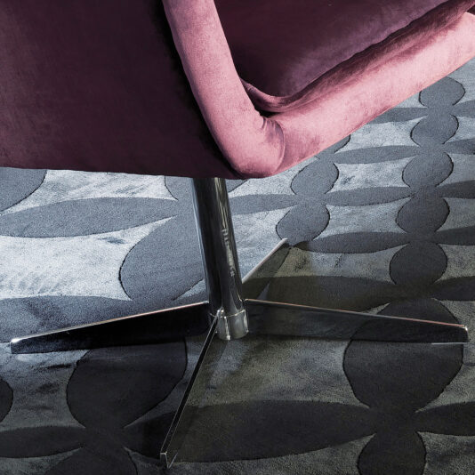 The Italian Button Upholstered Velvet Dining Chair, with its purple fabric seat and backrest on a metallic base, sits elegantly on a dark, patterned carpet.