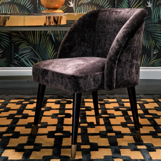 A Modern Designer Curved Velvet Dining Chair with dark wooden legs rests on a geometric-patterned rug, set against tropical wallpaper. A wooden table with a globe enhances the scene's eclectic style.