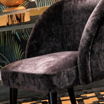Modern Designer Curved Velvet Dining Chair The Modern Designer Curved Velvet Dining Chair in plush dark gray, featuring sleek black legs, adds a contemporary touch to the room. It pairs beautifully with a wooden table and a wall decorated with elegant abstract plant designs for a stylish setup.