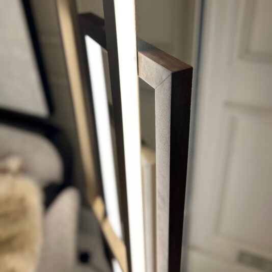 Close-up image of the edge of two picture frames leaning against a wall. The frames are different colors; one is light brown and the other is dark brown. An *Italian Geometric Designer Modern Floor Lamp* subtly illuminates the scene, with part of a cushioned chair draped in fur-like material visible in the background.