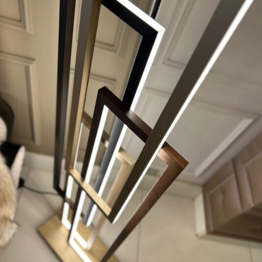 The Italian Geometric Designer Modern Floor Lamp, featuring intersecting rectangular frames in black, bronze, and gold illuminated along the edges, stands in a modern room with beige paneled walls and a light-colored floor.