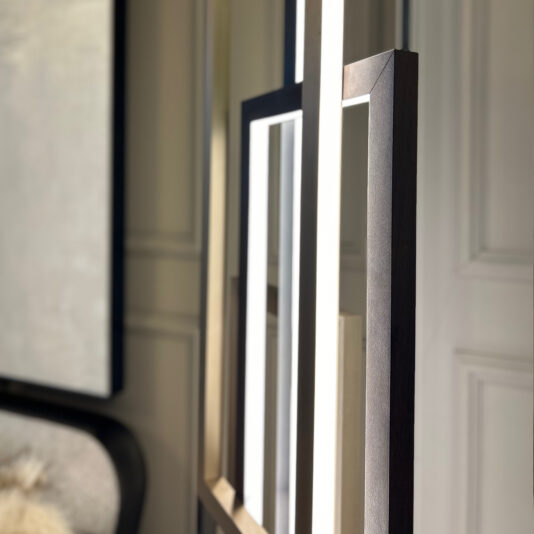 Close-up of the Italian Geometric Designer Modern Floor Lamp composed of rectangular frames with LED lighting. The lamp casts shadows on a paneled wall in a minimalist interior, revealing sleek dark and light-colored finishes on the furniture and other decor elements.