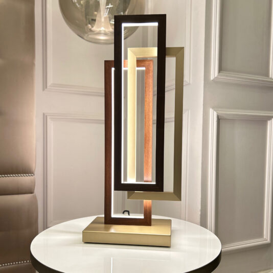 The Modern Italian Geometric Designer Table Lamp, showcasing a distinctive design with overlapping rectangular frames in wood and metallic finishes, emits a warm light. It is positioned on a white, round table set against a paneled wall. To the left, there's a partial view of a tufted chair.