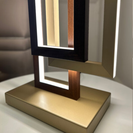 A Modern Italian Geometric Designer Table Lamp featuring interlocking rectangular frames made of wood and metal, illuminated by inner LED lights, is placed on a round white table. The background includes part of a gray upholstered chair, adding to the scene's geometric charm.