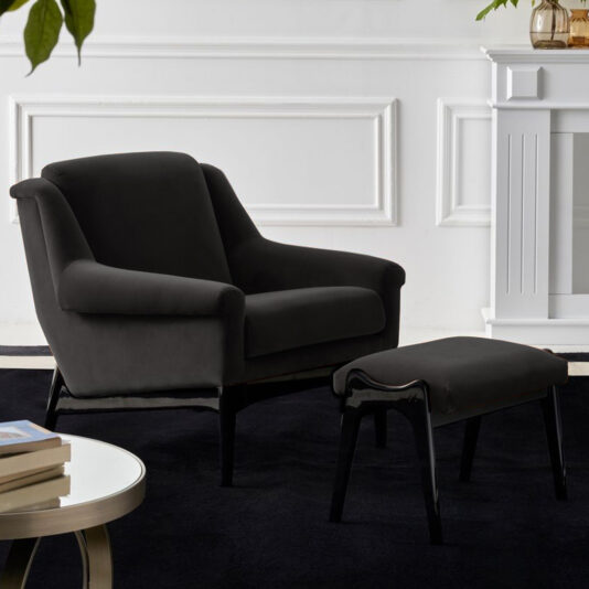 The Luxury High End Modern Black Velvet Armchair with a matching ottoman is placed on a dark rug in a chic room featuring white paneled walls, a decorative fireplace, and a round glass coffee table.