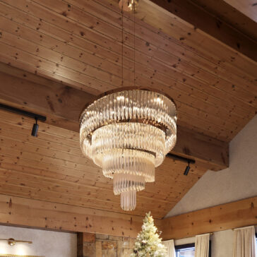 Gold Plated Spiral Crystal Designer Chandelier The Gold Plated Spiral Crystal Designer Chandelier hangs from a wooden ceiling with beams, its cascading glass elements shimmering. Below, a decorated Christmas tree stands near the corner of a room with rustic stone and wood walls.