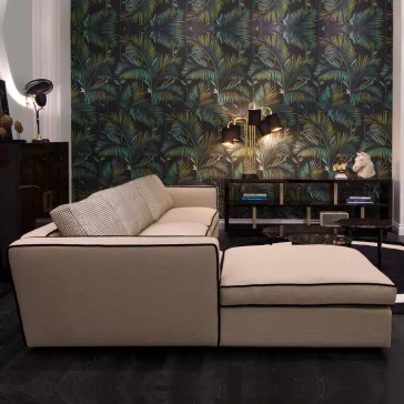 Italian Designer Modern Modular Corner Sofa An Italian Designer Modern Modular Corner Sofa in beige, featuring a chaise lounge, sits in front of dark tropical-themed wallpaper. Two sleek wall sconces provide lighting above a console table adorned with artistic decor and books. The room showcases a contemporary and stylish design complemented by dark wood flooring.