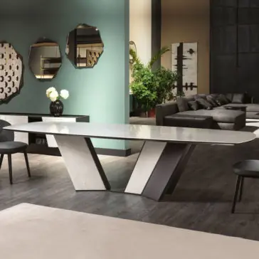 A modern dining room showcases a sleek Contemporary Rectangular Calacatta Dining Table on an angular base, encircled by black chairs. The backdrop features a green wall adorned with geometric mirrors, a sideboard displaying flowers, a large abstract painting, and a cozy seating area furnished with dark sofas and lush plants.