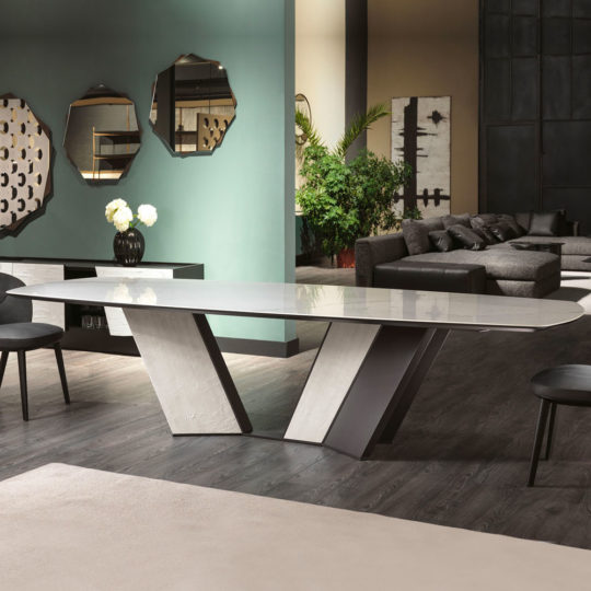 A modern dining room showcases a sleek Contemporary Rectangular Calacatta Dining Table on an angular base, encircled by black chairs. The backdrop features a green wall adorned with geometric mirrors, a sideboard displaying flowers, a large abstract painting, and a cozy seating area furnished with dark sofas and lush plants.