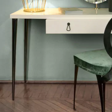 Italian Designer Ivory Lacquered Dressing Table With Mirror A modern workspace featuring the Italian Designer Ivory Lacquered Dressing Table With Mirror, which showcases a white desk with black tapered legs. Adorning the desk are a golden wire pen holder, a closed book, and an elegant decorative item. Positioned next to the dressing table is a green velvet chair. The entire setting is complemented by a wooden floor with a herringbone pattern.