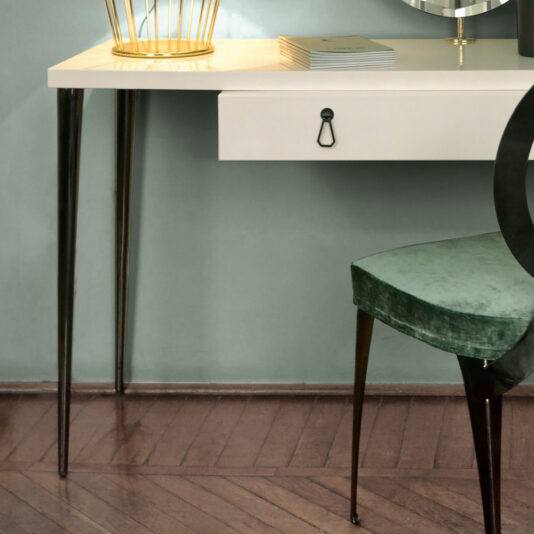 A modern workspace featuring the Italian Designer Ivory Lacquered Dressing Table With Mirror, which showcases a white desk with black tapered legs. Adorning the desk are a golden wire pen holder, a closed book, and an elegant decorative item. Positioned next to the dressing table is a green velvet chair. The entire setting is complemented by a wooden floor with a herringbone pattern.