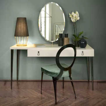 Italian Designer Ivory Lacquered Dressing Table With Mirror The Italian Designer Ivory Lacquered Dressing Table with Mirror, featuring slim black legs, is set against a muted green wall. Adorned with a round mirror, it also showcases a black table lamp, two bottles, a potted orchid, and a vase. Positioned in front is a green upholstered chair with a circular back.