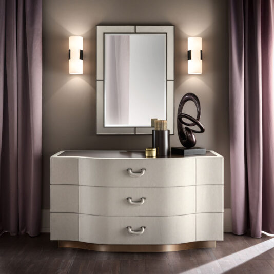 A sophisticated vanity setup showcasing a modern dresser with three curved drawers, adorned with décor pieces including a candle and a sculpture. The standout feature is the Italian Designer Leather Wall Mirror mounted above the dresser, flanked by two contemporary wall sconces.