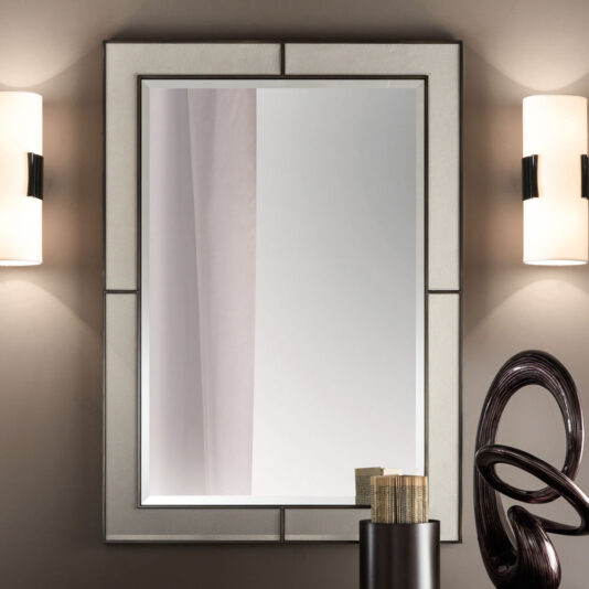 An Italian Designer Leather Wall Mirror with a geometric frame is flanked by two modern cylindrical wall sconces with white shades. Below the mirror, a contemporary sculpture stands next to a small stack of books on a table or shelf.
