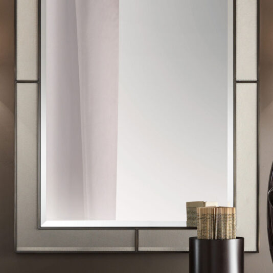 An Italian Designer Leather Wall Mirror with a stylish rectangular frame is mounted on a wall. The reflection shows part of a light-colored curtain. Below the mirror, there is a modern black vase containing several rolled-up paper tubes. The background wall has a neutral tone.