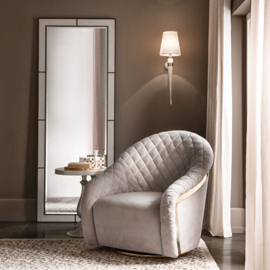 A cozy room features a gray, quilted armchair with a wooden frame next to a small, round side table adorned with decorative items. An Italian Designer Leather Wall Mirror leans stylishly against the wall, while a wall sconce with a white shade provides soft lighting. Light curtains frame the window.