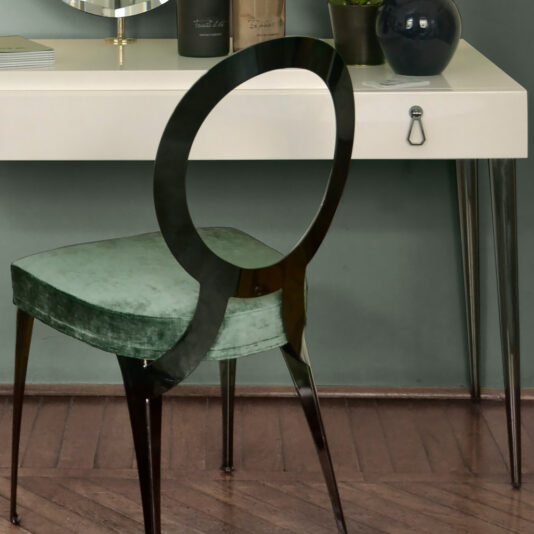 A modern workspace featuring an Italian Designer Ivory Lacquered Dressing Table with a drawer and silver tapered legs, exuding sleek sophistication. A stylish black chair with a circular backrest and green seat cushion sits at the desk, which is accented by a round mirror. The setup is completed with books and a potted plant for added charm.