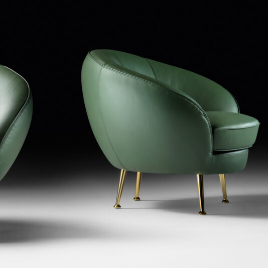 A stylish green, high-end Contemporary Designer Leather Occasional Armchair with a rounded back and seat, supported by sleek gold legs. The chair is positioned on a gray surface against a black background, showcasing its modern and elegant design. Only a part of a second chair is visible on the left side.