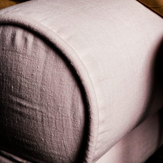 Close-up of a beige upholstered cylindrical cushion on the Contemporary Retro Designer Linen Sofa, likely serving as an armrest. The fabric appears soft with visible stitching and texture. The lighting casts subtle shadows that highlight the fabric's contours and details, enhancing its retro charm.