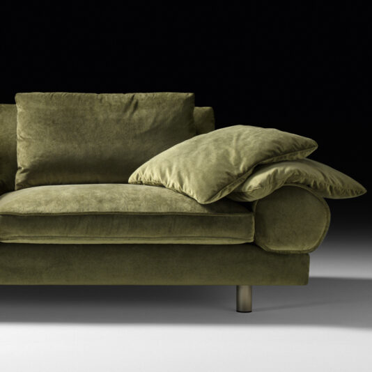 Contemporary Retro Inspired Designer Sofa A close-up of the Contemporary Retro Inspired Designer Sofa in luxurious green velvet against a black background. The focus is on the armrest with its layered cushions and cylindrical base, complemented by slightly visible metallic legs. This contemporary piece boasts a textured and plush appearance.