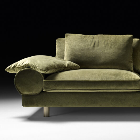 Contemporary Retro Inspired Designer Sofa A Contemporary Retro Inspired Designer Sofa in olive green velvet is partially shown against a black background. The focus is on one side, featuring a cushion on the armrest and a metallic sofa leg visible beneath. The soft, luxurious texture exemplifies its contemporary designer appeal.