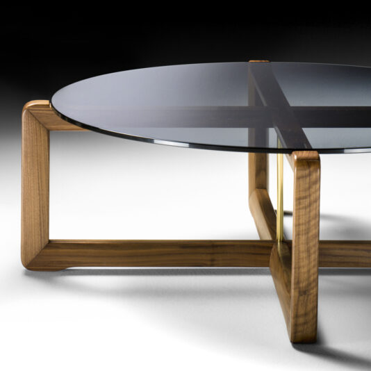 Contemporary Walnut Round Glass Coffee Table The Contemporary Walnut Round Glass Coffee Table features a circular glass top and a wooden base. The wooden base is designed with intersecting geometric legs, offering a stylish and clean appearance. This round glass table is elegantly displayed on a white surface against a black background.