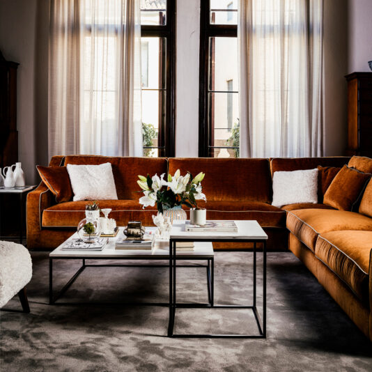 Designer Marble Contemporary Square Coffee Table A stylish living room featuring a plush rust-colored sectional sofa with white accent pillows and throws, a pair of Designer Marble Contemporary Square Coffee Tables with elegant marble tops, and a vase of white flowers. Sheer curtains allow natural light to filter through large windows, illuminating the space.